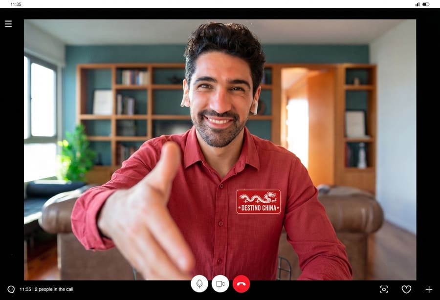 Captura de pantalla de una videollamada donde un hombre sonriente con camisa roja extiende su mano hacia la cámara en un saludo. En su camisa lleva un logotipo que dice "Destino China".